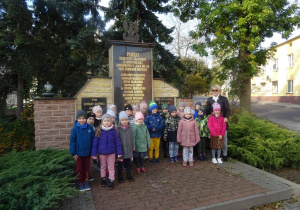 Grupa dzieci z panią Arletą stoi pod pomnikiem poległych podczas II wojny światowej.